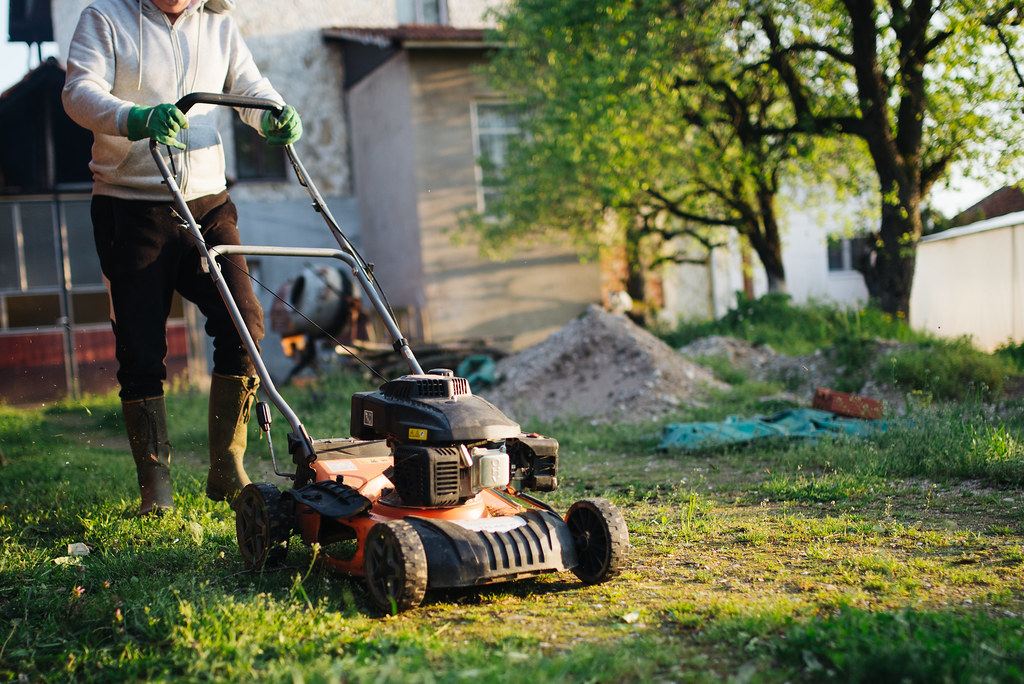 A landscaper mowing a backyard lawn beside garden beds